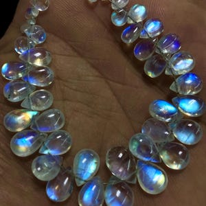 May include: A necklace of teardrop-shaped moonstone beads. The translucent stones have a shimmering play of colour, with flashes of blue, purple, and yellow. The beads are strung on a thin, silver-coloured wire, creating a delicate and elegant appearance.