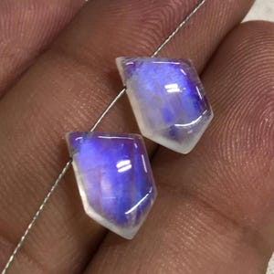 May include: Two iridescent moonstone gemstones, shield-shaped. The stones show white, blue, and purple hues, with a polished, reflective surface. A thin, silver wire runs through the stones, suggesting they are for jewellery making. Each stone is approximately 1 cm in length.