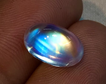 May include: A polished, oval-shaped gemstone with a translucent appearance. The stone displays a play of color, with iridescent flashes of blue, yellow, and white. The gem is held between fingers, showcasing its size and luster.