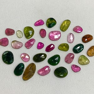 May include: A collection of 25 faceted gemstones in various shades of green, pink, and brown. The gemstones are arranged on a white surface.
