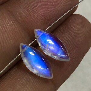 May include: Two marquise-cut moonstone beads with a vibrant blue flash. The gemstones are strung on a thin, silver wire, showcasing their iridescent quality. The beads are held against a neutral background, highlighting their colour and shape.