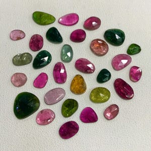 May include: Assortment of colorful gemstones in various shapes and sizes, including pink, green, and yellow. The gemstones are arranged on a white surface.