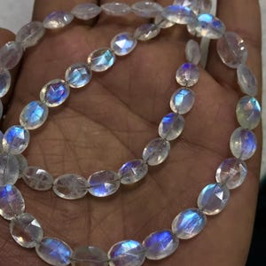 May include: A strand of oval-shaped moonstone beads with iridescent blue and white colours. The beads are strung on a clear thread.
