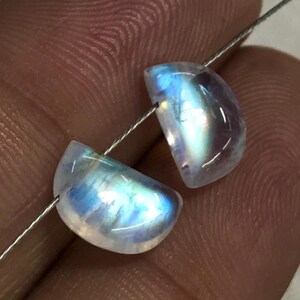 May include: Two polished, half-moon shaped moonstone beads strung on a thin wire. The beads display a mix of translucent white, blue, and iridescent colours. The gemstones are smooth and have a subtle sheen, ideal for jewellery making.
