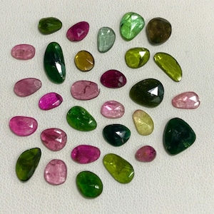 May include: Assortment of green, pink, and red gemstones in various shapes and sizes. The gemstones are arranged in a heart shape on a white surface.