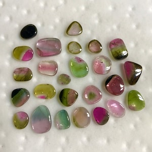 May include: A collection of 20+ green, pink, and black gemstones. The gemstones are various shapes and sizes, and have a smooth, polished finish.