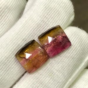 May include: Two faceted, square-cut gemstones. The gems display a gradient of colors, transitioning from a dark golden-brown at the top to a vibrant pink at the base. The stones are set against a white fabric background.