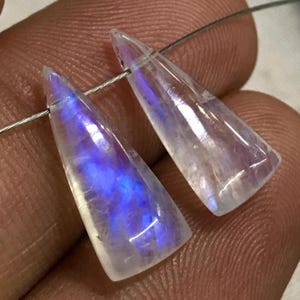 May include: Two iridescent rainbow moonstone teardrop shaped pendants with a pointed tip. The stones are translucent and have a white base with blue and purple iridescence.