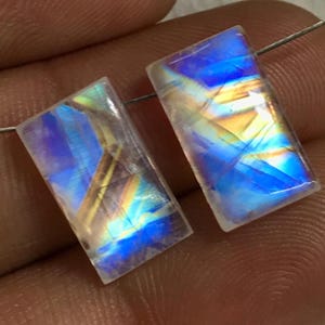 May include: Two iridescent rainbow moonstone cabochons, cut in a rectangular shape. The stones have a translucent white base with flashes of blue, green, and gold.