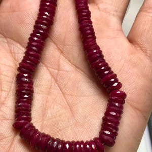 May include: A strand of deep red ruby beads, each bead is faceted and has a slightly rounded shape. The beads are strung on a clear thread and are approximately 1/2 inch in diameter.