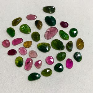 May include: A collection of green, pink, and brown gemstones. The gemstones are various shapes and sizes, and have a smooth, polished finish.