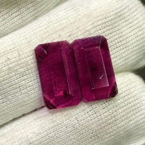 May include: Two rectangular, faceted, deep magenta gemstones are displayed on a white, textured surface. The gems have a transparent appearance with visible internal structures and sharp edges, suggesting they are cut and polished.