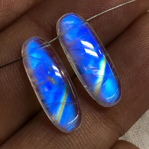 May include: Two oval-shaped rainbow moonstone cabochons with iridescent blue, purple, and gold colours. The stones are translucent and have a smooth, polished surface.