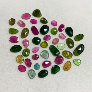 May include: A collection of green, pink, and red gemstones, cut into various shapes and sizes. The gemstones are arranged on a white surface.