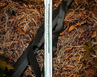 May include: A silver sword with a decorative handle and etched markings on the blade, resting on a bed of dry leaves and grass. A black leather sheath and strap are positioned beside the sword.