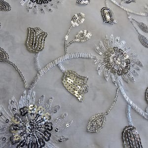 May include: Silver sequined fabric with a floral design. The fabric features a repeating pattern of flowers and leaves, all embellished with silver sequins.