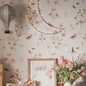 Può includere: Una cameretta con carta da parati floreale e decorazioni a farfalla. Una lampada a forma di luna crescente e un'illustrazione per bambini incorniciata sono presenti. Una lampada a farfalla rosa e un vaso di tulipani rosa completano la scena.