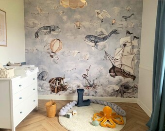 May include: A nursery room with a blue and white wallpaper featuring whales, ships, and hot air balloons. The room has a white dresser with blue knobs, a wicker basket, and a plush octopus toy.