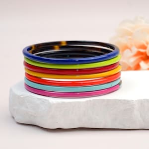 May include: A stack of eight thin, circular bangle bracelets in a variety of solid colours including dark tortoise shell, royal blue, lime green, red, yellow, orange, turquoise, and magenta, arranged in a vertical column on a white surface.