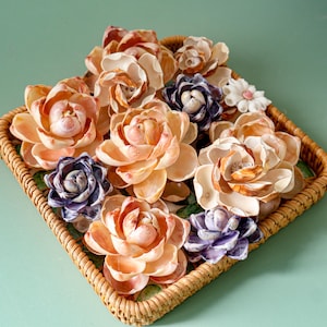 Puede incluir: Un conjunto de flores hechas con conchas marinas en tonos melocotón, crema y morado, dispuestas en una cesta cuadrada de mimbre. Las flores tienen detalles intrincados en los pétalos y están sobre un fondo verde claro. La cesta es de material tejido de color natural.