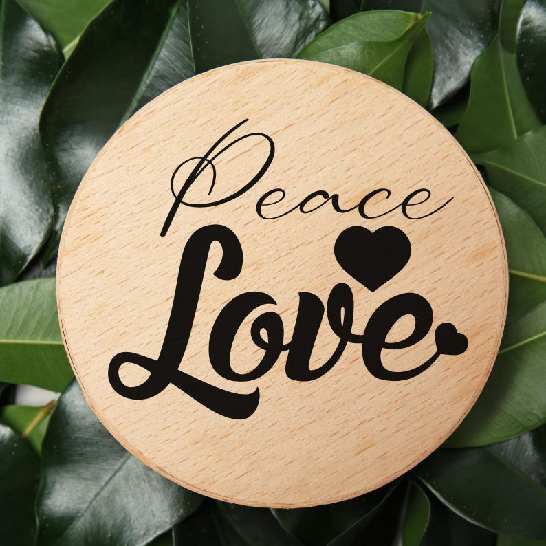 May include: A circular wooden coaster featuring the words "Peace Love" in black script, with a black heart. The coaster is set against a backdrop of green leaves.