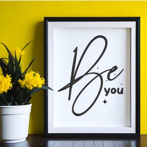 May include: A framed print featuring the words "Be you" in black script. The artwork is set against a white background within a black frame. A potted plant with yellow flowers is positioned to the left of the frame against a yellow wall.
