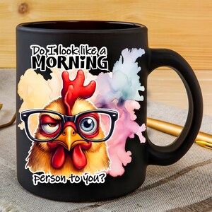Pode incluir: Uma caneca de cer&acirc;mica preta fosca com um gr&aacute;fico colorido de uma galinha usando &oacute;culos de aro preto. O texto diz: Do I look like a MORNING person to you? A galinha tem uma crista e barbelas vermelhas, com um olho semicerrado e outro aberto.