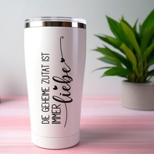 May include: A white stainless steel tumbler with a clear lid, featuring black text that reads DIE GEHEIME ZUTAT IST IMMER liebe with small heart icons. The design is printed vertically on the side of the cylindrical container.