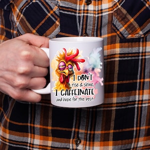 Pode incluir: Uma caneca de cer&acirc;mica branca com uma ilustra&ccedil;&atilde;o colorida de uma galinha despenteada e sonolenta com penas bagun&ccedil;adas. O texto diz: I DON'T rise & shine, I CAFFEINATE and hope for the best. O design inclui salpicos de cor estilo aguarela.