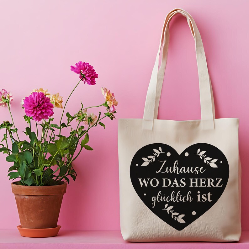 May include: A cream-colored canvas tote bag featuring a large black heart graphic on the front. The heart contains the German text "Zuhause WO DAS HERZ gl&uuml;cklich ist" surrounded by small white leaf and dot accents, with two long shoulder straps.