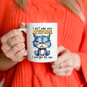 May include: White ceramic mug featuring a grumpy blue cat illustration with the text "I don't need your ATTITUDE, I brought my own." The cat has an angry expression with furrowed eyebrows. The mug has a standard handle.