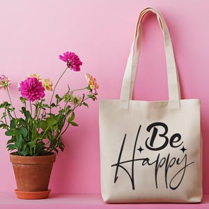 May include: A cream tote bag with the words "Be Happy" in black script. The bag has long handles and is set against a pink background. A potted plant with pink flowers is to the left.