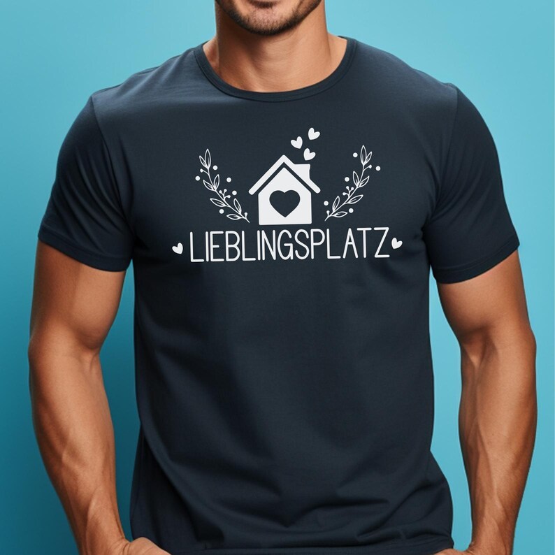 May include: A dark navy blue short-sleeved t-shirt featuring a white graphic print of a house with a heart in the center, surrounded by small hearts and leafy branches, with the text "LIEBLINGSPLATZ" printed in white capital letters below.