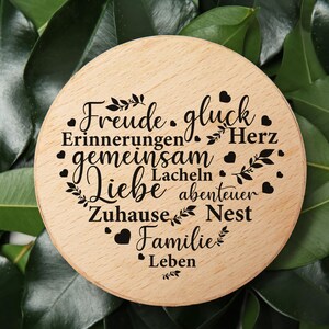 May include: A circular light-colored wooden plaque featuring black laser-engraved German words arranged in a heart shape, including Freude, gluck, Erinnerungen, Herz, gemeinsam, Lacheln, Liebe, abenteuer, Zuhause, Nest, Familie, and Leben with small hearts.