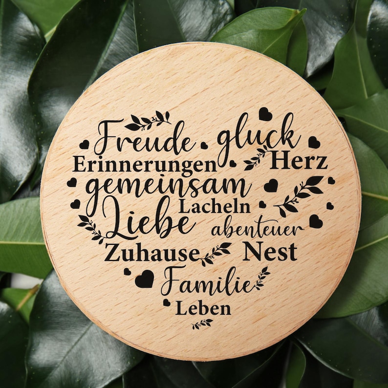 May include: A circular light-colored wooden plaque featuring black laser-engraved German words arranged in a heart shape, including Freude, gluck, Erinnerungen, Herz, gemeinsam, Lacheln, Liebe, abenteuer, Zuhause, Nest, Familie, and Leben with small hearts.