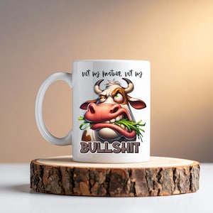 May include: White ceramic mug with a cartoon illustration of a cow with an angry expression, eating grass. The mug has the text "not my pasture, not my BULLSHIT". The mug is on a wooden coaster.