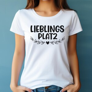 May include: A white short-sleeved t-shirt featuring the black text LIEBLINGS PLAT2 printed on the chest. Below the text is a decorative graphic of two symmetrical leafy branches meeting at a small solid black heart in the center.