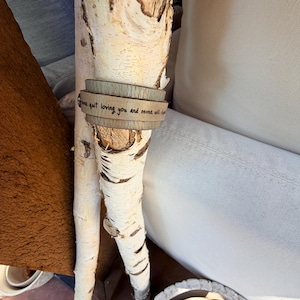 May include: A light brown leather bracelet wrapped around a birch branch. The bracelet has the inscription "I will never quit loving you and never will". The birch branch is white with dark markings.