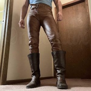 May include: Brown leather trousers featuring a high-waisted design with a double-buckle belt closure at the waist. The trousers are paired with dark brown leather mid-calf boots that have a smooth finish and a rounded toe construction.