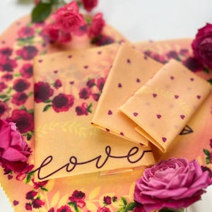 May include: A set of three reusable food wraps with a floral pattern and the word "love" written on the top wrap. The wraps are made of beeswax, jojoba oil, and tree resin. The wraps are a sustainable alternative to plastic wrap.