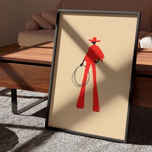May include: Framed art print featuring a minimalist illustration of a figure in red attire, holding a lasso. The figure is wearing a hat and standing against a beige background. The artwork is in a black frame, leaning against a wooden table.