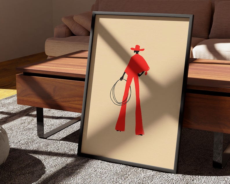 May include: Framed art print featuring a minimalist illustration of a figure in red attire, holding a lasso. The figure is wearing a hat and standing against a beige background. The artwork is in a black frame, leaning against a wooden table.