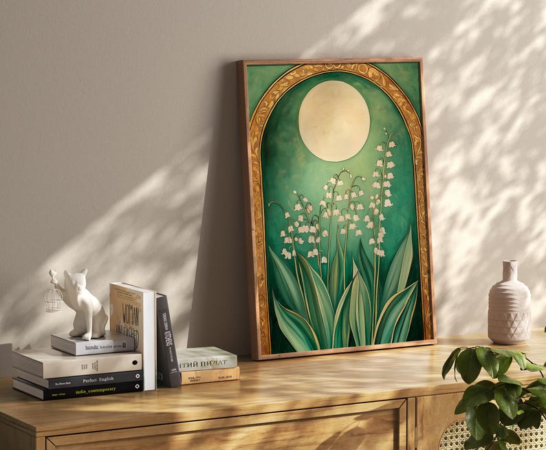 May include: A framed art print featuring a botanical illustration of lily of the valley flowers. The artwork has a green and gold color scheme, with a large moon in the background. The print is displayed on a wooden cabinet with books and decorative items.