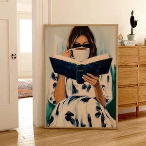 May include: A framed art print depicts a person reading a book and drinking from a white mug. The person is wearing a white dress with blue floral patterns. The artwork is in a light wood frame and rests on a wooden floor.