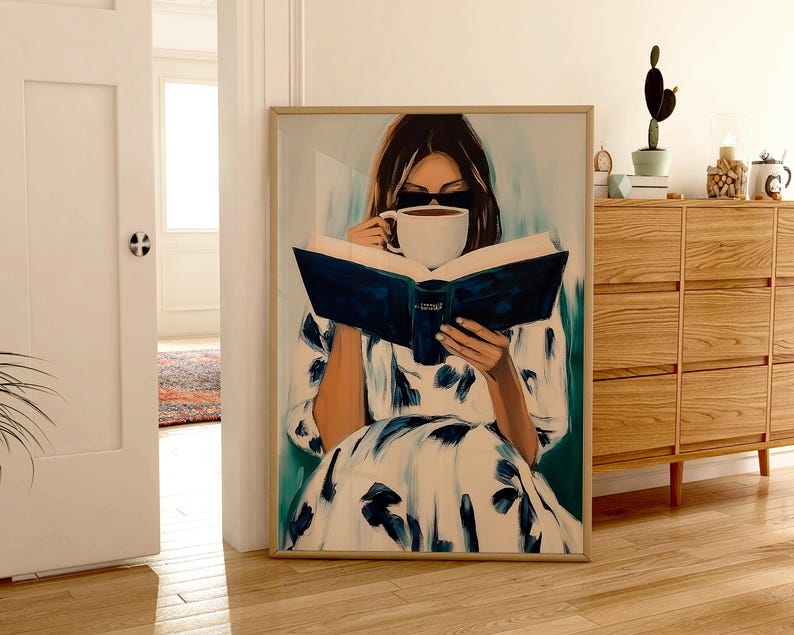May include: A framed art print depicts a person reading a book and drinking from a white mug. The person is wearing a white dress with blue floral patterns. The artwork is in a light wood frame and rests on a wooden floor.
