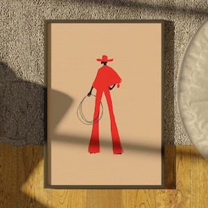 May include: Framed art print featuring a stylized illustration of a person in red cowboy attire, holding a lasso. The figure is set against a beige background, with a dark frame. The artwork evokes a Western theme.