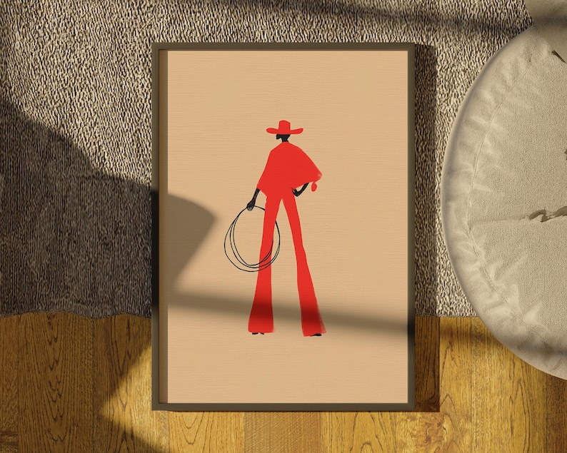 May include: Framed art print featuring a stylized illustration of a person in red cowboy attire, holding a lasso. The figure is set against a beige background, with a dark frame. The artwork evokes a Western theme.