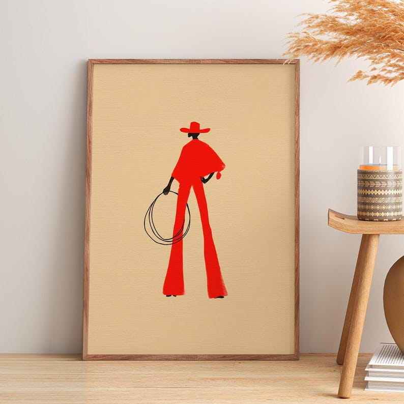 May include: Framed art print featuring a stylized illustration of a figure in a red outfit, holding a lasso. The figure is wearing a hat and cape, with flared pants. The background is a light beige color.