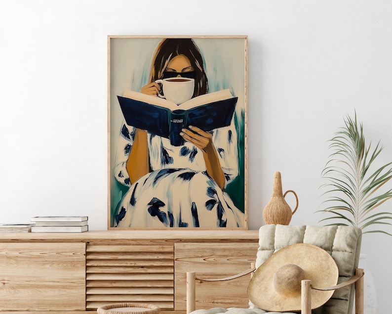 May include: A framed art print depicts a person wearing sunglasses, reading a book, and drinking from a white mug. The person is wearing a white dress with blue floral patterns. The book has a dark blue cover.
