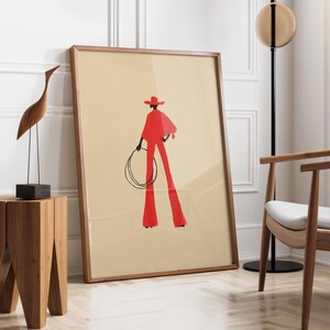May include: A framed art print featuring a stylized illustration of a person in a red outfit holding a lasso. The artwork has a beige background and a wooden frame. A wooden sculpture of a bird and a wooden side table are in the foreground.